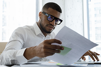 Serious African American manager in stylish glasses reading corporate document with note, sales report, boss order, doing paperwork. Legal expert checking document at workplace with laptop.