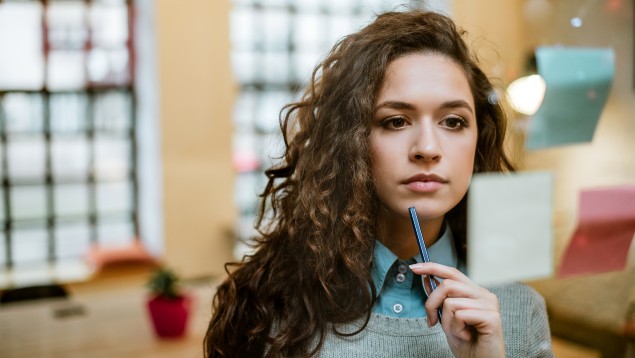 A person contemplates while holding a pen near their chin, looking at sticky notes on a glass wall in a brightly lit office setting.