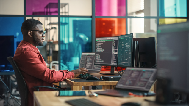 A person working intently at a desk with multiple monitors displaying code and data analysis, in an office with colorful glass partitions.