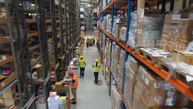 Workers wearing safety vests and helmets inside a large warehouse with tall shelves stocked with various items.
